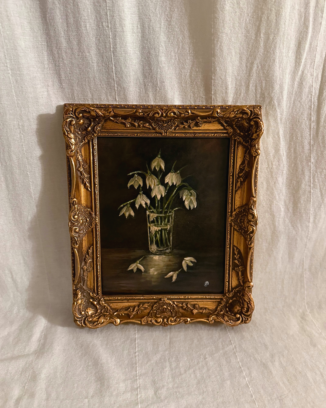 Framed painting of snowdrops in a glass vase with a dark moody backdrop photographed on a textured white background