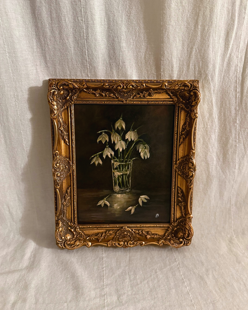 Framed painting of snowdrops in a glass vase with a dark moody backdrop photographed on a textured white background