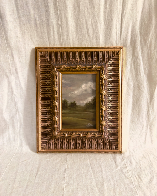 Wide ornate antique gilt frame with a grey small moody british countryside landscape painting inside photographed on a textured white background